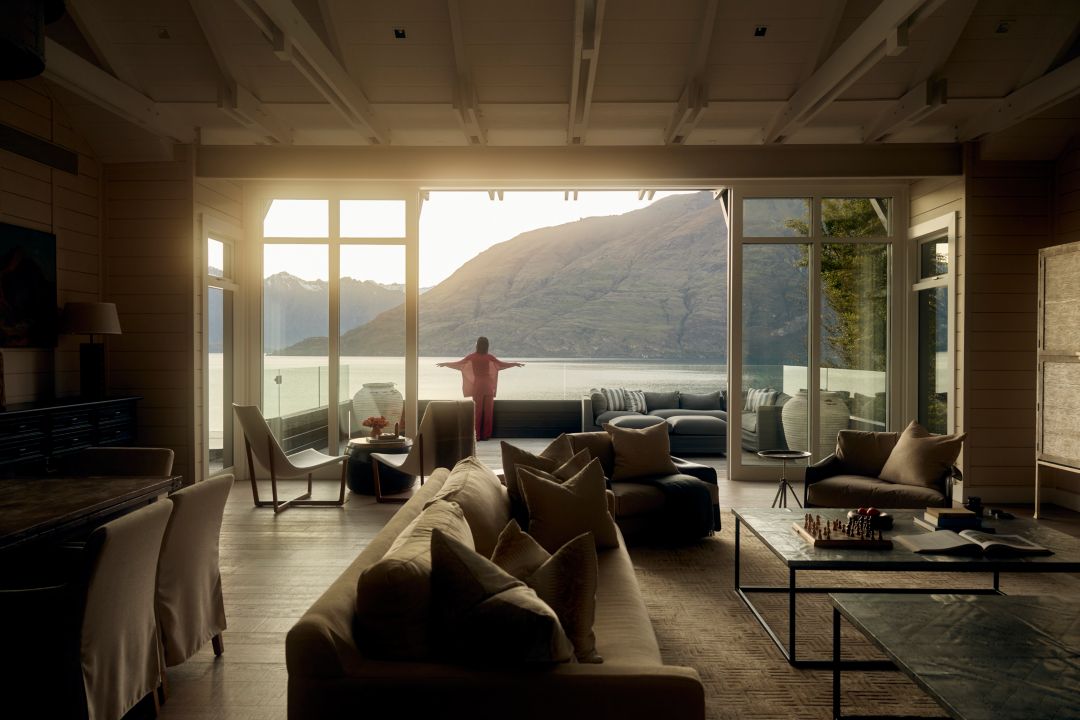 The living and dining room in a luxury villa with neutral furnishing and open doors leading out to a private balcony where a person is standing overlooking Lake Wakatipu and the surrounding mountains. 